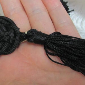 Peut inclure: Un pompon noir avec un détail de nœud décoratif. Le pompon est fait de fils noirs étroitement tissés, et le nœud est un design rond et complexe. Le pompon est tenu dans une main sur un fond blanc.