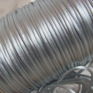 May include: A spool of light gray satin cord. The cord is tightly wound, revealing a smooth, shiny texture. The image shows the cord in a close-up view, highlighting its delicate appearance. Ideal for crafting and jewelry making.