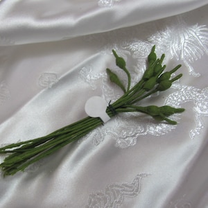 May include: A bundle of small, green artificial flower buds tied together with green wire and a white tag. The stems are long and thin, and the buds are a darker shade of green. The background is a white, patterned fabric.