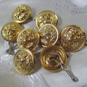 May include: A collection of gold-colored metal buttons with intricate embossed designs. The buttons are round and feature detailed relief patterns. Each button has a small metal loop on the back for attachment. The buttons are arranged on a white, textured surface.