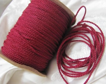 May include: A spool of burgundy-colored rope sits next to a length of the same rope, coiled. The rope is tightly woven and appears to be made of a soft material. The spool is made of wood.