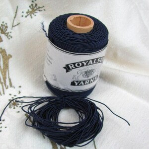May include: A spool of navy blue yarn with a wooden core and a label that reads "ROYALS YARNS." A separate pile of navy blue yarn is next to the spool. The yarn is on a white fabric with a subtle floral pattern.