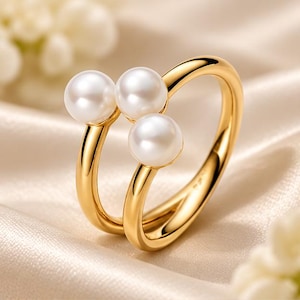 May include: A gold ring featuring three round white pearls arranged in a cluster. The band is smooth and polished, with an open design that allows the pearls to sit prominently at the top. The metal has a bright, reflective yellow gold finish.