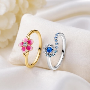 Puede incluir: Un anillo dorado con una flor de esmalte rosa y una piedra central transparente, y un anillo plateado con un diseño en espiral engastado con múltiples piedras azules de varios tonos, ambos expuestos sobre una tela blanca suave.