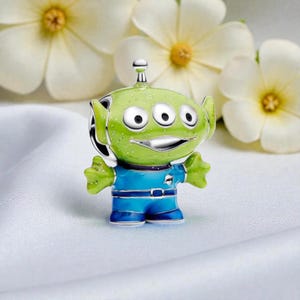 Peut inclure: Un charm argenté en forme de personnage extraterrestre à trois yeux. La figurine présente une tête vert vif avec des accents scintillants, trois yeux blancs et un large sourire. Elle porte un uniforme bleu avec une ceinture et des bordures métalliques.