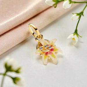 May include: A gold charm shaped like a lily flower with white and yellow petals, a pink centre, and small clear stones embedded in the middle. The charm features a gold loop at the top for attachment to a bracelet or necklace chain.