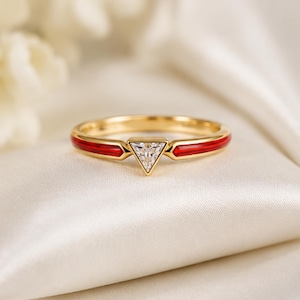 Könnte beinhalten: Ein goldener Ring mit einem zentralen, dreieckigen, klaren Edelstein, der von zwei horizontalen roten Emaileinlagen am Ringband flankiert wird. Das Metallband ist poliert und glatt, was eine klare geometrische Ästhetik mit einem Kontrast erzeugt.