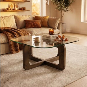 Puede incluir: Una mesa de centro cuadrada con tapa de cristal transparente y base de madera marrón oscuro con diseño en forma de cruz. Sobre la mesa hay una pequeña taza de cerámica, dos libros apilados, una vela encendida y una rama con hojas de otoño.