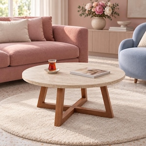 Op de afbeelding: Een ronde salontafel met een beige gespikkeld stenen blad en een bruin houten kruisvormig onderstel. Op de tafel staat een klein glas met donkere thee op een schoteltje en een gesloten boek met een lichte kaft. Het ontwerp heeft een rond blad.