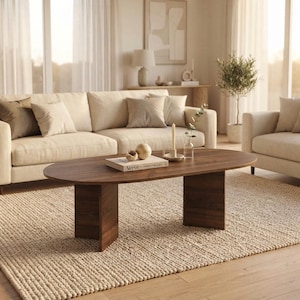 May include: An oval coffee table made of dark brown wood with two wide, flat vertical supports. On the table surface sits a book titled Verve, a small wooden sphere, and a tall, thin white candle in a holder with a small glass vase of greenery.
