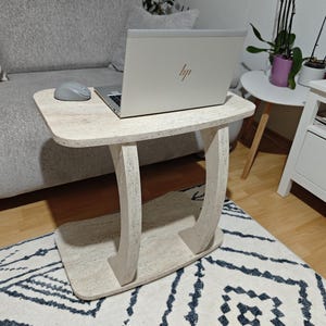 Può includere: Un tavolino color beige chiaro maculato con piano e base rettangolari collegati da due supporti curvi ad arco. Un computer portatile argentato con logo HP visibile e un mouse grigio sono posizionati sulla superficie del tavolino.