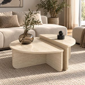 May include: A set of two nested coffee tables made of beige travertine stone with a natural porous texture. The tables feature semi-circular tops and vertical slab legs. A dark glass vase with green branches and a black stone sphere sit on top.