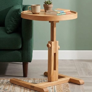 Peut inclure: Une table d'appoint en bois clair avec un plateau rond et un mécanisme de réglage en hauteur comprenant une colonne de support verticale centrale, un bouton circulaire de réglage et une base en H large et stable au sol.