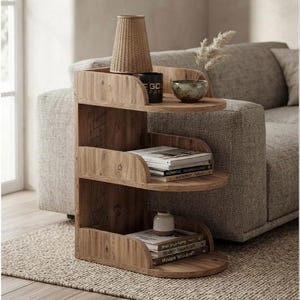 Puede incluir: Una mesa auxiliar de madera de tres niveles con un diseño curvo y abierto. El estante superior sostiene una lámpara tejida, una taza negra con el texto "GOLD FARM" y un cuenco pequeño. Los estantes intermedio e inferior contienen libros y un jarrón.
