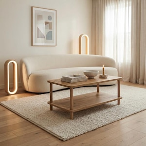 May include: A rectangular wooden coffee table with a lower shelf, positioned in front of a curved cream-coloured sofa. The table holds a stack of books, a small bowl, and a lit candle on a stone base. Two oval-shaped floor lamps emit a warm glow.