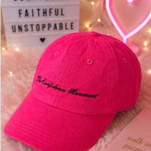 May include: A bright hot pink baseball cap featuring the embroidered black text The Confidence Movement on the front panel. The cap has a structured crown, a curved brim, and stitched eyelets for ventilation, showcasing a vibrant solid color design.