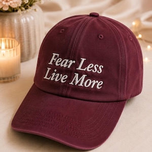 May include: A deep burgundy baseball cap featuring the embroidered white text "Fear Less Live More" on the front panel. The cap has a structured crown, a curved brim with visible stitching, and a button at the top center of the fabric dome.