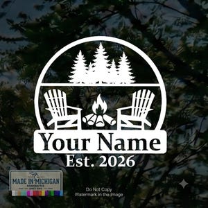 Op de afbeelding: Een witte cirkelvormige sticker met een silhouet van dennenbomen boven twee Adirondack-stoelen en een kampvuur. Onder de stoelen bevindt zich een rechthoekige banner met de tekst Your Name en Est. 2026. Een klein label vermeldt Made in Michigan.