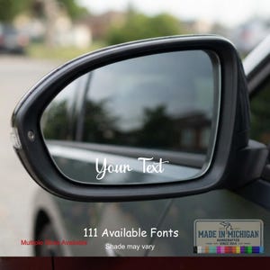 May include: A black car side mirror featuring a white custom vinyl decal with the script text Your Text applied to the glass surface. The mirror housing is dark and sleek, reflecting the blurred outdoor environment in the background.