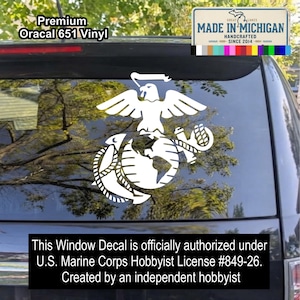 May include: A white vinyl decal of the United States Marine Corps emblem featuring an eagle, globe, and anchor applied to a vehicle rear window. Text includes Premium Oracal 651 Vinyl, Made in Michigan, and licensing information.