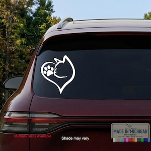 May include: A white vinyl decal applied to the rear window of a maroon vehicle. The decal features a stylized heart shape formed by the silhouette of a cat head on the right side and a paw print on the left side, creating a cohesive graphic design.