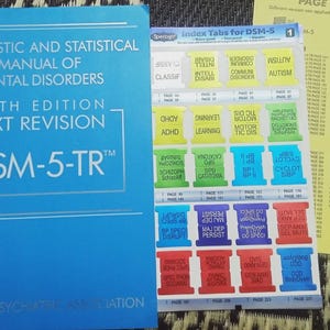 May include: A bright blue Diagnostic and Statistical Manual of Mental Disorders Fifth Edition Text Revision DSM-5-TR book next to a sheet of colorful adhesive index tabs and a yellow reference page with a printed page table.