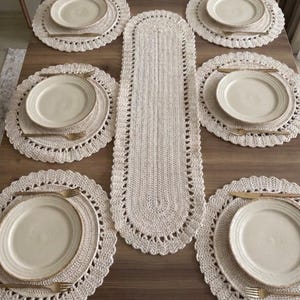 May include: A set of six round crocheted placemats and one long oval crocheted table runner in an off-white color. Each placemat is topped with a plain cream-colored ceramic plate and a gold-toned fork, creating a coordinated dining table arrangement.