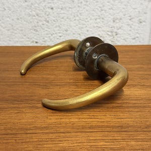 May include: A pair of brass door handles featuring curved levers and circular mounting plates. The metal has a warm, golden-toned finish with visible signs of age and patina. The handles are connected by a central mechanism with screw holes.