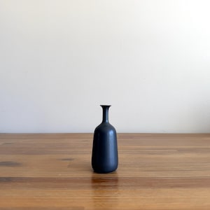 Könnte beinhalten: Eine kleine, mattschwarze Knospenvase mit einem schlanken, länglichen Hals und einem abgerundeten, zylindrischen Körper. Die Vase hat eine glatte, gleichmäßige dunkle Oberfläche und eine schmale Öffnung oben und steht auf einem stabilen Boden.