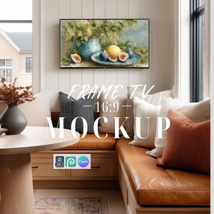 May include: A digital television screen displaying a still life painting of a blue pitcher, a yellow lemon, and sliced figs on a plate. The screen is mounted on a white paneled wall above a tan leather bench with various throw pillows.