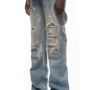 Op de afbeelding: Lichtblauwe spijkerbroek van denim met een versleten afwerking, meerdere scheuren, gerafelde randen en witte verfspatten. De broek heeft een rechte snit en een doorleefde uitstraling, waarbij stukken stof zichtbaar zijn onder de gescheurde delen.