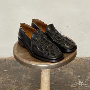 Può includere: Un paio di mocassini in pelle nera caratterizzati da una superficie superiore testurizzata e arricciata con dettagli di cucitura visibili. Le scarpe presentano una punta quadrata, un tacco basso e una fodera interna liscia color cuoio.