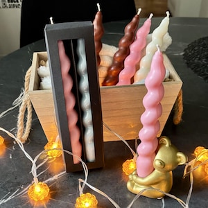 May include: A collection of spiral-shaped taper candles in shades of pink, white, and brown. Some candles are displayed in a black rectangular box with a window, while others are held in a gold-coloured bear-shaped candle holder and a wooden crate.