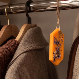 Könnte beinhalten: Eine sechseckige orangefarbene Wachstafel mit eingebetteten getrockneten blauen Blütenblättern, die an einer dünnen braunen Schnur an einer Metallkleiderstange hängt. Die Wachstafel hat eine glatte Oberfläche und hängt vor verschiedenen Kleidungsstücken.