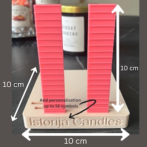 Puede incluir: Un soporte cuadrado beige de 10 cm por 10 cm con dos pilares verticales rosas acanalados. El borde frontal presenta el texto en relieve "Istorija Candles" y una flecha que apunta a la base con el texto "Add personalisation up to 16 symbols".