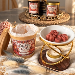 May include: A wax warmer set featuring a gold metal frame, a white ceramic dish holding red leaf and heart-shaped wax melts, and a small tea light candle. Beside it sits a cylindrical cardboard container labeled Falling Leaves with a wooden lid.