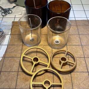 Op de afbeelding: Drie goudkleurige, ronde kunststof lontcentreerhulpmiddelen met een ontwerp met drie spaken, geplaatst voor twee doorzichtige glazen kaarsenpotten en twee amberkleurige glazen potten, elk met een witte lont op een metalen voetje.