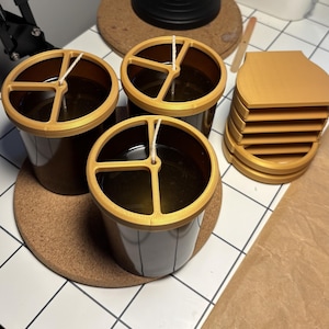 May include: Three dark brown cylindrical candle containers filled with wax, each topped with a gold-coloured plastic wick centring tool. Beside the candles sits a stack of four matching gold-coloured plastic components with a flat, rounded top edge.
