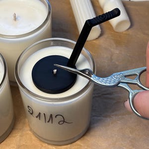 May include: A clear glass candle container filled with white wax, featuring a black circular wick centering tool and a black angled straw-like accessory. A silver stork-shaped scissor is trimming the wick. The glass has the word "enlive" written on it.