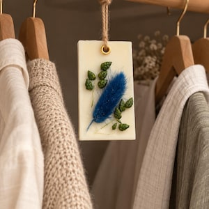 Può includere: Una tavoletta di cera rettangolare color bianco sporco appesa a un bastone appendiabiti in legno tramite un laccio in corda di juta. La tavoletta presenta un pennacchio blu di erba essiccata, piccole foglie verdi e un occhiello metallico dorato.