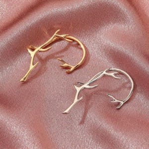 May include: Two ear cuffs, one in a polished gold tone and one in a polished silver tone. Each piece features a curved, open design that mimics the organic, branching structure of a tree twig or antler, with small, pointed protrusions along the frame.