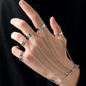 May include: A silver-toned hand harness featuring five plain metal rings connected by delicate link chains that extend from each finger to a central wrist chain. The jewellery piece is constructed from polished metal links and smooth bands.