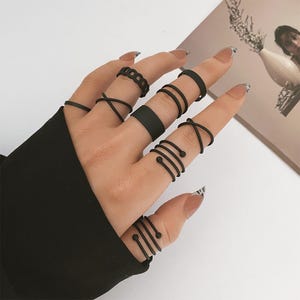 May include: A collection of ten matte black rings worn on the fingers. The set includes various designs such as thin bands, a wide band, crisscross patterns, and spiral wraps with small spherical ends, all featuring a consistent dark finish.