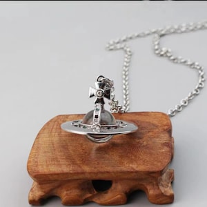 May include: A silver-toned necklace featuring a spherical pendant with a surrounding ring and a cross topper. The pendant is embellished with small clear crystals and a central dark orb. The chain is a classic silver-toned link style.
