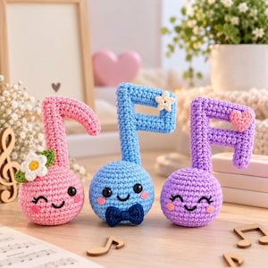 May include: Three crocheted musical note figures in pink, blue, and purple. The pink note features a white flower, the blue note has a dark blue bow tie and a small star, and the purple note displays a pink heart. Each has a smiling face.