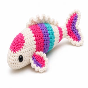Könnte beinhalten: Ein gehäkeltes Fischspielzeug mit einem Streifenmuster in Creme, Pink, Lila und Türkis. Der Fisch hat ein einzelnes schwarzes Kunststoffauge sowie eine Rückenflosse, eine Brustflosse und eine Schwanzflosse mit cremefarbenem Wellenrand.