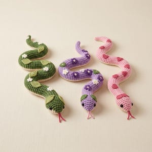 Op de afbeelding: Drie gehaakte slangen in groen, paars en roze. De groene slang is versierd met witte bloemen en groene blaadjes. De paarse slang heeft paarse en witte bloemen. De roze slang is gedecoreerd met rode hartvormen. Elke slang heeft een roze tong.