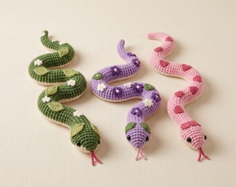 May include: Three crocheted snake toys in green, purple, and pink. The green snake features white flowers and green leaves. The purple snake has purple and white flowers. The pink snake is decorated with red heart shapes. Each has a pink tongue.