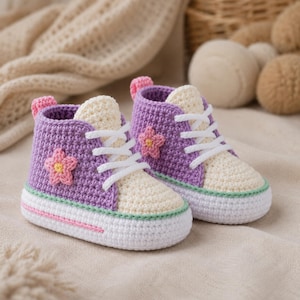 Peut inclure: Une paire de baskets pour bébé crochetées à la main en fil violet et crème. Chaque chaussure présente des lacets blancs, une applique fleur rose sur le côté, un liseré vert clair au-dessus de la semelle blanche et une rayure rose.