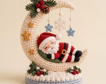 May include: A crochet ornament featuring a cream-colored crescent moon base with a sleeping Santa Claus figure in a red and white suit. The moon is decorated with green pine sprigs, red berries, a brown pinecone, and hanging blue stars and white snowflakes.